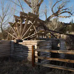 An old wooden waterwheel is situated behind a log fence, with its spokes and paddles visible.