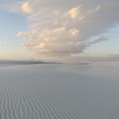 An expanse of rippled white sand stretches out into the distance, meeting a cloudy blue sky at the horizon.