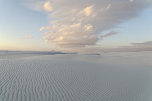 An expanse of rippled white sand stretches out into the distance, meeting a cloudy blue sky at the horizon.