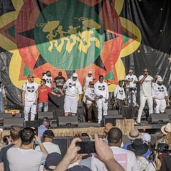 A group of men wearing white t-shirts with black logos are standing on stage in front of a large colorful banner.