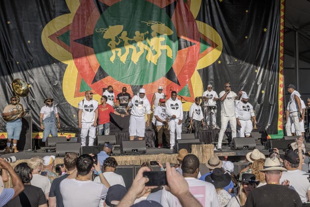 A group of men wearing white t-shirts with black logos are standing on stage in front of a large colorful banner.