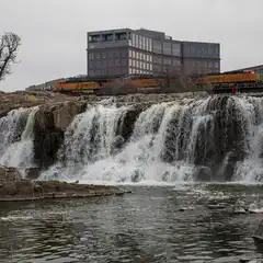 A waterfall cascades over a rocky ledge into a pool of water. An orange train is on tracks above the waterfall, with multi-story buildings in the background.