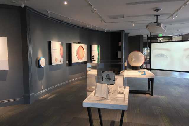 A room with a white table holding an exhibit about vision, surrounded by posters on the walls and a large screen displaying images of eyes.