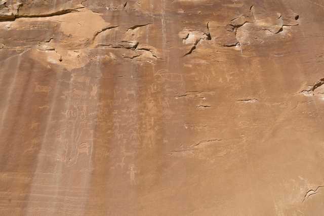 A massive sandstone cliff face features numerous petroglyphs that depict various animals and human-like figures.