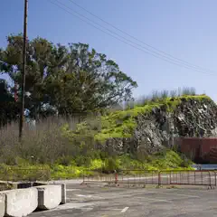 A rocky hill is covered in green vegetation and is bordered by a parking lot and a red metal fence.