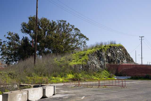 A rocky hill is covered in green vegetation and is bordered by a parking lot and a red metal fence.