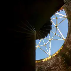 A spiral staircase leading upwards into an open, circular space with a curved ceiling and exposed structural beams. The walls are made of brick.