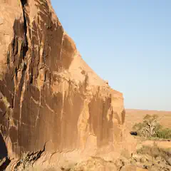 A desert landscape features a prominent rocky cliff face, set against a backdrop of scrubby vegetation and a clear blue sky.