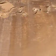 A massive sandstone cliff face features numerous petroglyphs that depict various animals and human-like figures.