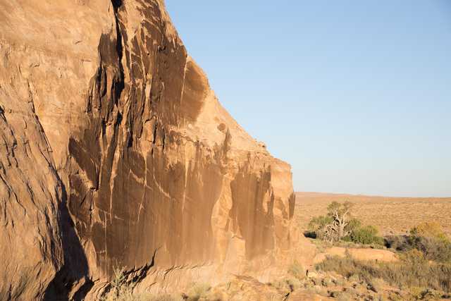 A desert landscape features a prominent rocky cliff face, set against a backdrop of scrubby vegetation and a clear blue sky.
