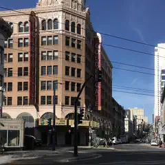 An intersection in a city's theater district, featuring a prominent building with a sign that reads 'Now Playing'.