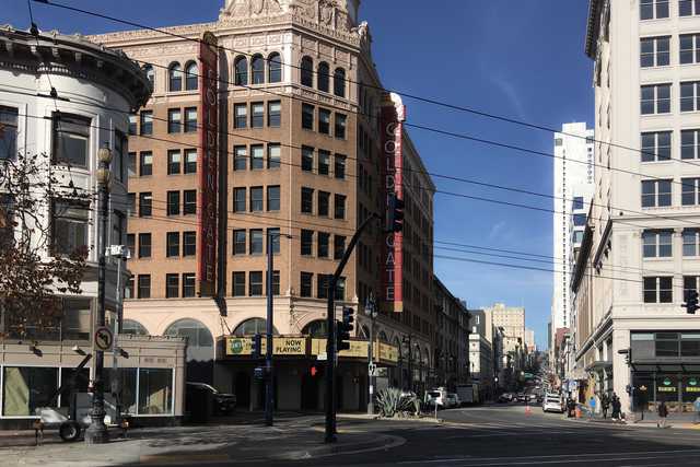 An intersection in a city's theater district, featuring a prominent building with a sign that reads 'Now Playing'.