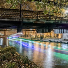 A river flows under a bridge at night, its surface reflecting vibrant lights that resemble neon trails or car headlights.