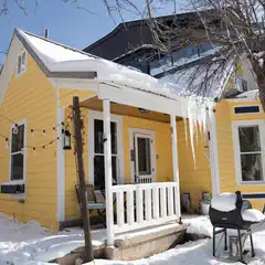 Snow covers the ground outside a small yellow house with white trim, featuring a front porch with a snow-covered railing and roof.