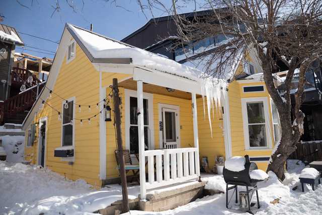 Snow covers the ground outside a small yellow house with white trim, featuring a front porch with a snow-covered railing and roof.