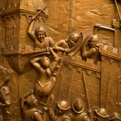 A wooden relief depicts a medieval siege, with soldiers scaling a castle wall while defenders on the ramparts wield weapons and shields.