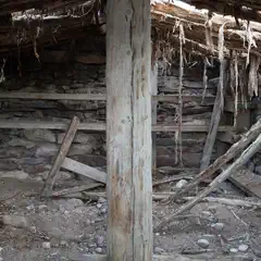 A dilapidated stone and wood structure with collapsed roof and walls, showing exposed wooden beams and debris scattered within.