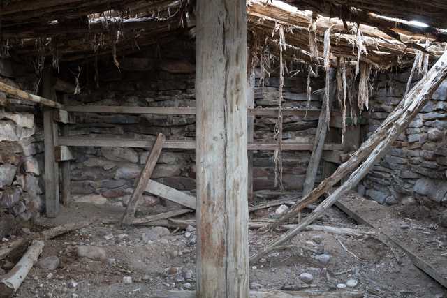 A dilapidated stone and wood structure with collapsed roof and walls, showing exposed wooden beams and debris scattered within.