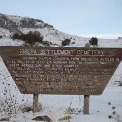 A wooden sign with information about a historical cemetery sits next to snow-covered ground and mountains in the background.
