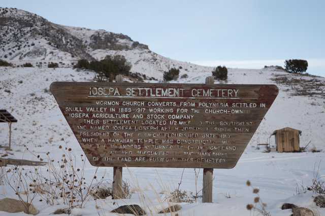 A wooden sign with information about a historical cemetery sits next to snow-covered ground and mountains in the background.