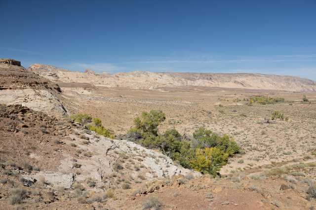 A desert landscape featuring rocky outcrops and scrubby vegetation stretches towards distant mountains under a clear blue sky.