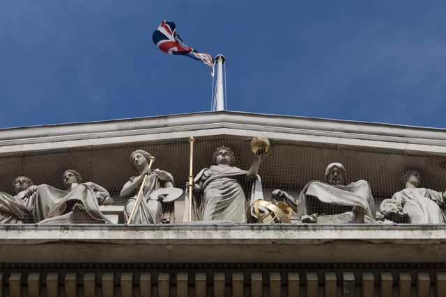 A Union Jack flies above a frieze of classical stone statues on a building facade.