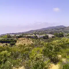 A view of a hillside covered with green shrubs, with a valley and houses visible below under clear skies.