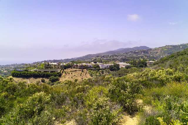 A view of a hillside covered with green shrubs, with a valley and houses visible below under clear skies.