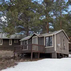 A small brown wooden cabin with a gray door and white trim is situated on a snowy ground, surrounded by trees.