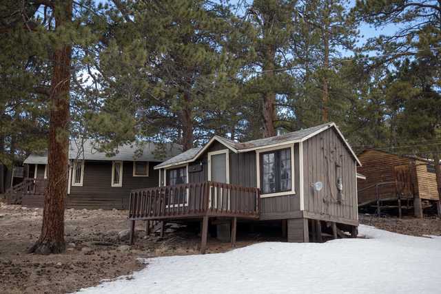 A small brown wooden cabin with a gray door and white trim is situated on a snowy ground, surrounded by trees.