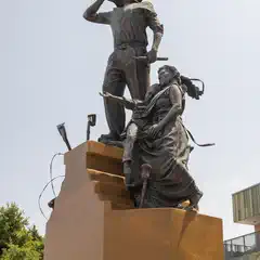 A statue of a man and woman, possibly depicting a historical event or figures, atop a pedestal with trees in the background.