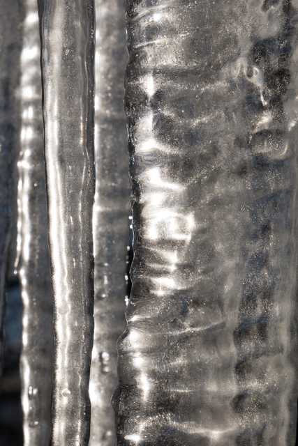 A close-up of icicles hanging from a structure, exhibiting intricate patterns and glimmering with light reflected off their surfaces.