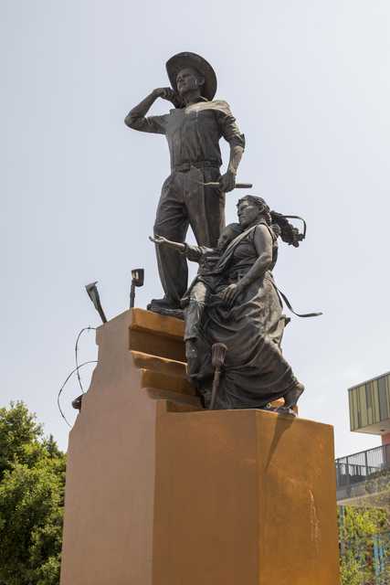 A statue of a man and woman, possibly depicting a historical event or figures, atop a pedestal with trees in the background.