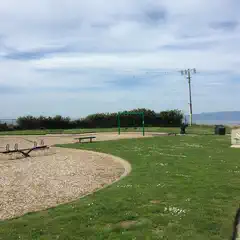 A playground with a seesaw and swing set sits on wood chips, adjacent to a paved path and a grassy area with a sign.