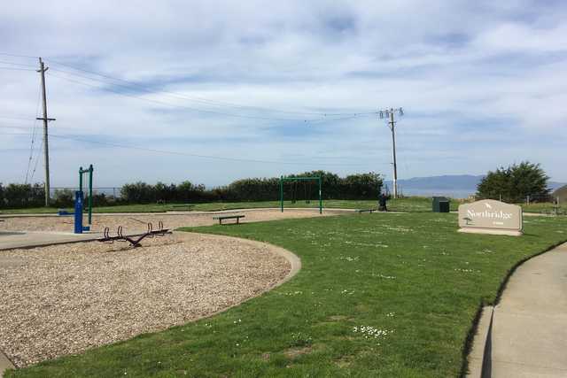 A playground with a seesaw and swing set sits on wood chips, adjacent to a paved path and a grassy area with a sign.