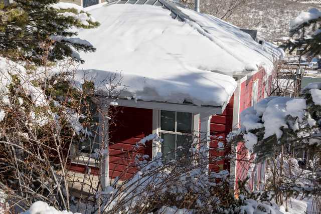 A red house with white trim and a snow-covered roof is visible through trees and bushes.