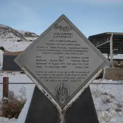A stone plaque with engraved text stands on a snowy ground near a fence and mountains in the background.