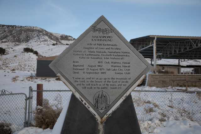 A stone plaque with engraved text stands on a snowy ground near a fence and mountains in the background.