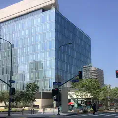 A large, modern building with many windows and glass panels, situated on a city street corner with traffic lights and pedestrians visible.