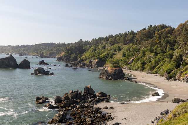 A sandy beach curves along a coastline with rock formations and a forested hillside.