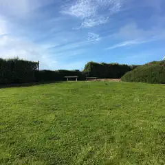 A grassy field under a blue sky with clouds, bordered by neatly trimmed hedges and a white house in the background.