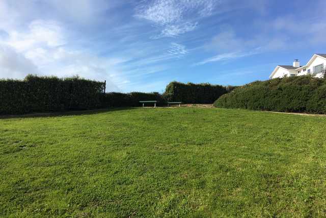 A grassy field under a blue sky with clouds, bordered by neatly trimmed hedges and a white house in the background.