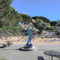 A playground features geometric climbing structures and a blue abstract sculpture on a paved surface.