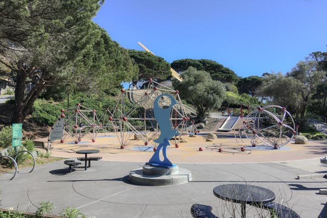 A playground features geometric climbing structures and a blue abstract sculpture on a paved surface.