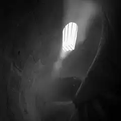 A narrow, spiraling staircase descends toward a barred window, with light streaming through the opening.