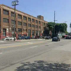 A long road with parked cars on the side. A brick building in the background. Trees along the sidewalk. It's a sunny day.