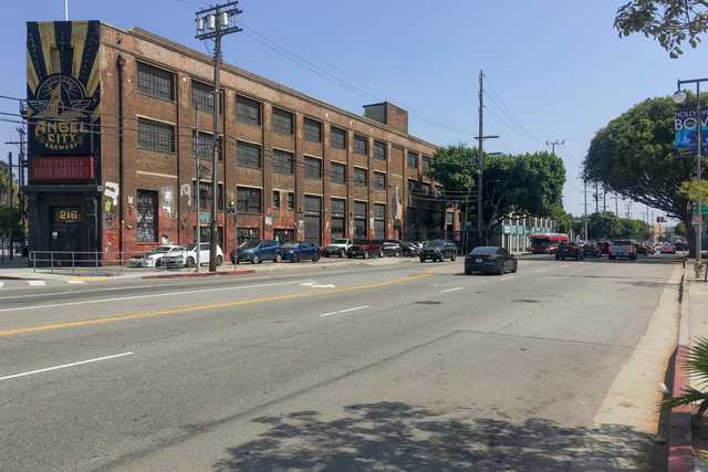 A long road with parked cars on the side. A brick building in the background. Trees along the sidewalk. It's a sunny day.