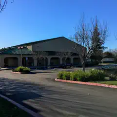 A building with a flat roof and an American flag on a pole in front of it, surrounded by trees and parked cars, under a clear blue sky.