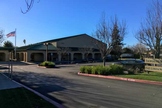 A building with a flat roof and an American flag on a pole in front of it, surrounded by trees and parked cars, under a clear blue sky.