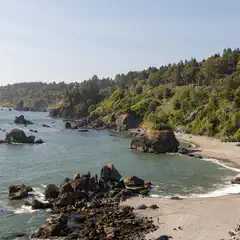 A sandy beach curves along a coastline with rock formations and a forested hillside.